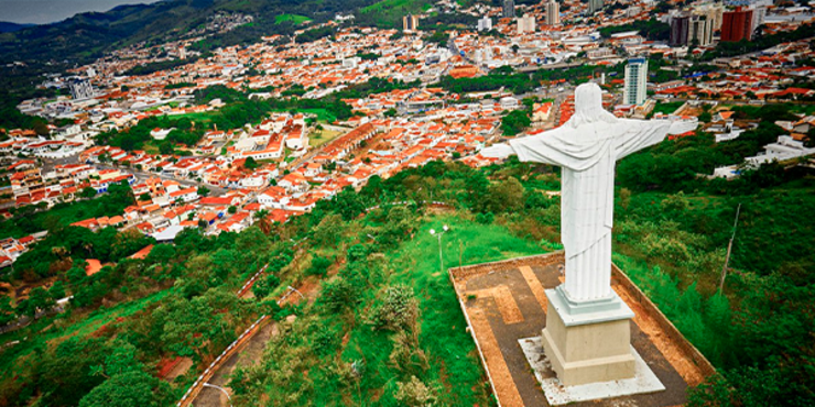 Vista de um cristo redentor branco no topo de um morro, de braços abertos para a cidade de Amparo, com muitas casas e alguns prédios isolados entre elas, e cercada por morros e vegetação. 