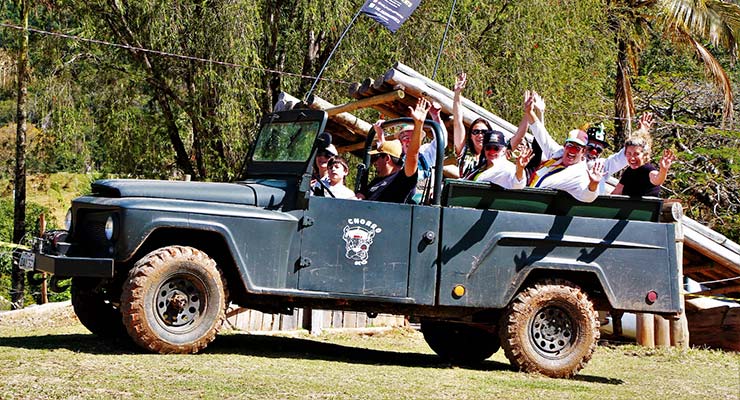 Grupo de pessoas sorrindo e acenando em um jipe off-road em terreno acidentado, aproveitando uma aventura ao ar livre.