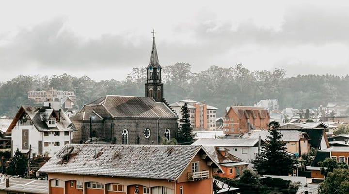 Imagem de uma cidade com prédios residenciais e uma igreja com torre, sob céu nublado, em um dia de inverno ou clima chuvoso.