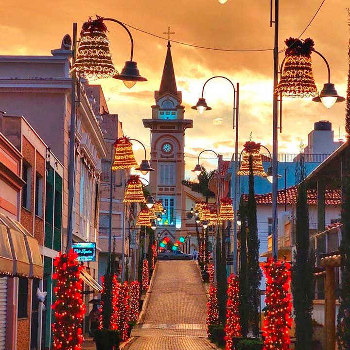 Rua decorada para o Natal com luzes, árvores decoradas com luzes vermelhas, iluminação natalina, prédios coloridos e uma torre com relógio ao fundo.
