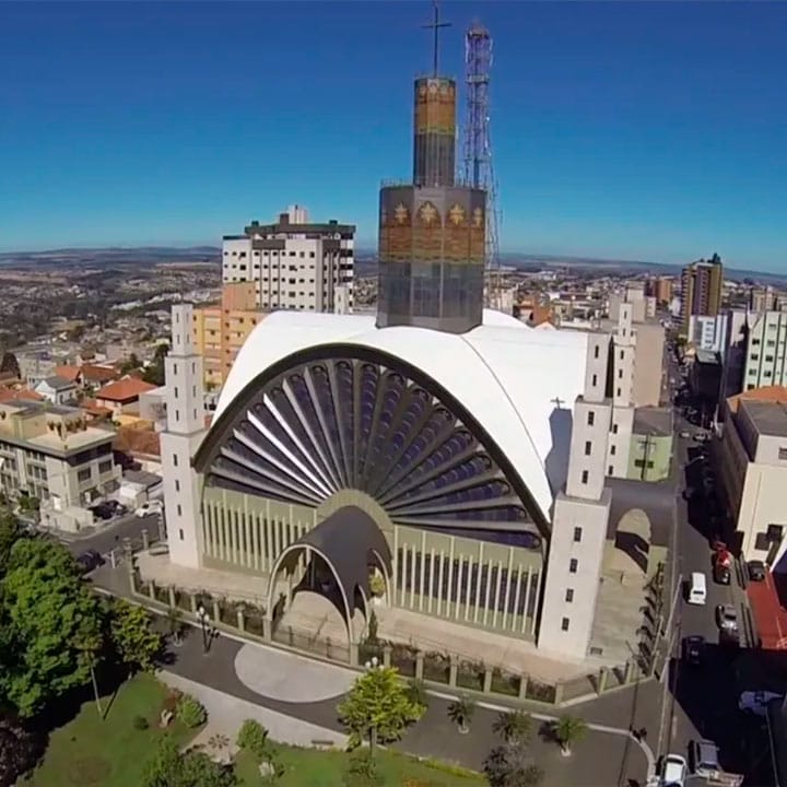 Vista aérea de uma igreja moderna e imponente na cidade, com uma estrutura arquitetônica única, cercada por edifícios e ruas movimentadas ao redor.