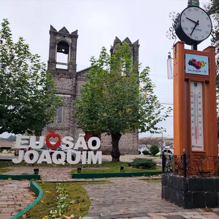 Imagem de uma praça em São Joaquim com uma igreja histórica ao fundo, uma árvore central e uma estação de medição de temperatura, promovendo turismo e cultura local.