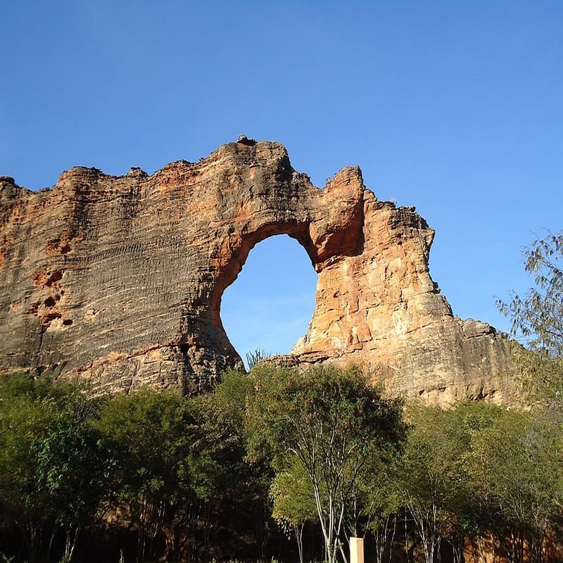 Formação rochosa com um grande buraco no meio, rodeada por vegetação e céu azul no fundo.