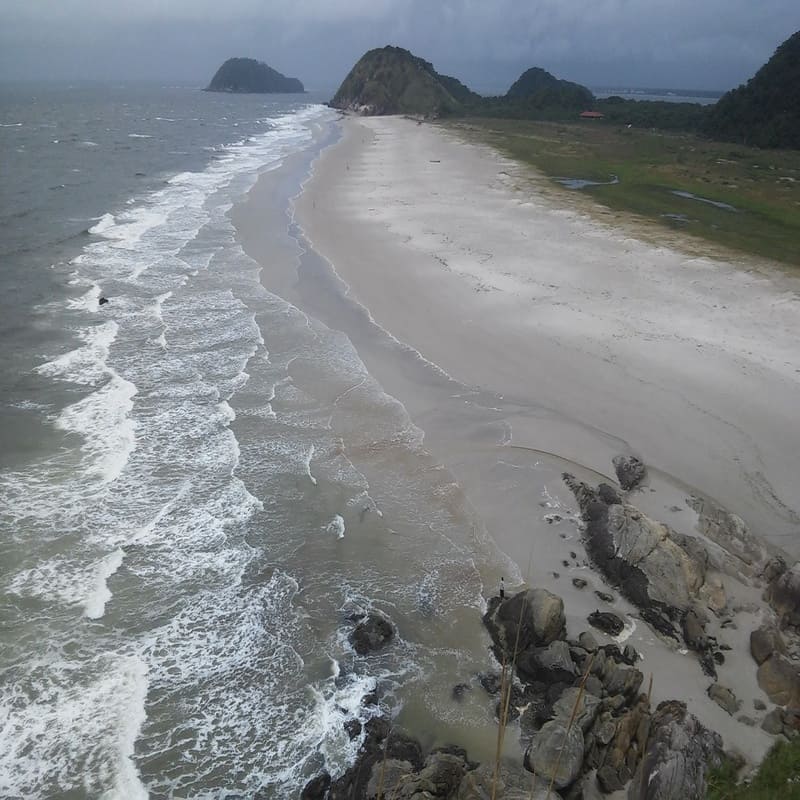 Abeira de uma praia deserta com ondas quebrando na areia e formações rochosas, cenário natural com dunas e morros ao fundo, sob céu nublado.