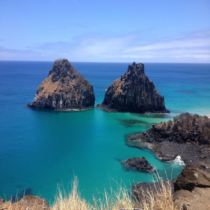Paisagem costeira com duas grandes rochas no mar azul, vista de ilha brasileira, destaque para beleza natural e águas cristalinas.
