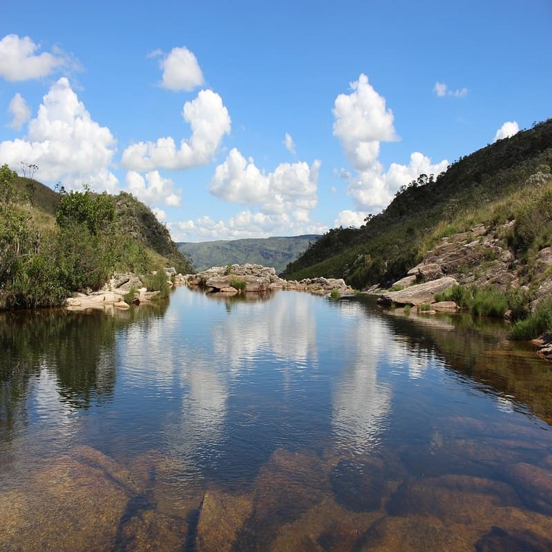 Paisagem natural de rio com vegetação e montanhas ao fundo sob céu azul com nuvens brancas, refletindo na água calma.